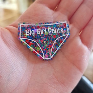 May include: A colourful glitter resin keychain shaped like underwear with the text "Big Girl Pants" in white.