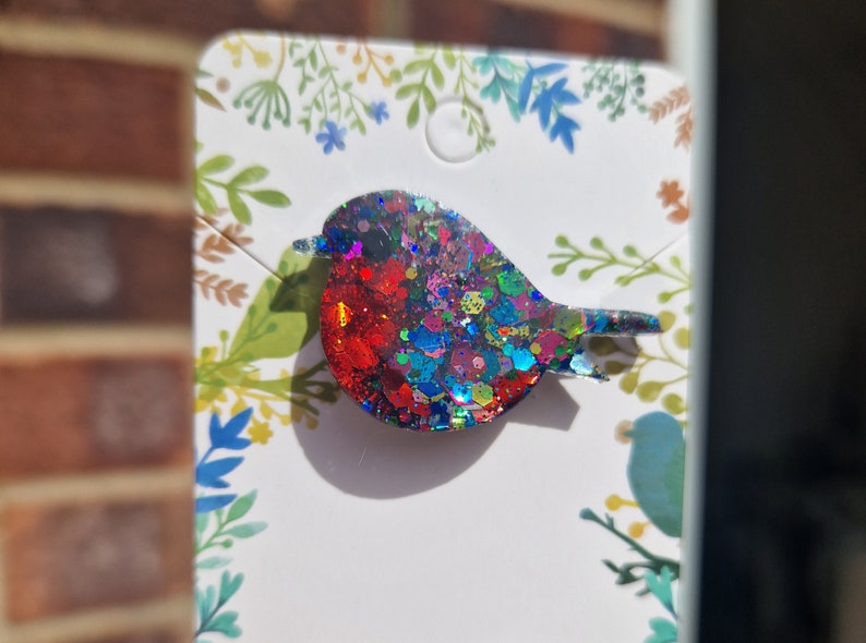 Peut inclure: Une broche en forme de rouge-gorge en paillettes rouges, bleues et vertes. Le rouge-gorge est en r&eacute;sine et a une finition brillante et iris&eacute;e.