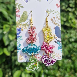 May include: Dangle earrings featuring three colorful dinosaur shapes in red, blue, green, yellow, and purple. The earrings have gold-colored hooks and are attached to a decorative card with floral and bird designs. The dinosaurs have glitter accents.