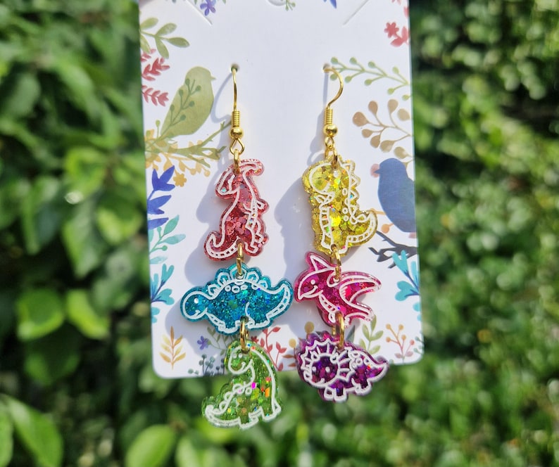 May include: Dangle earrings featuring three colorful dinosaur shapes in red, blue, green, yellow, and purple. The earrings have gold-colored hooks and are attached to a decorative card with floral and bird designs. The dinosaurs have glitter accents.