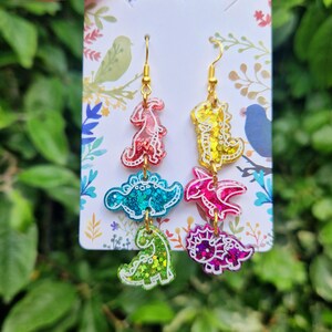 May include: Colorful dinosaur earrings with gold-colored hooks. Each earring features three dinosaur shapes in red, blue, green, yellow, pink, and purple with glitter accents. The earrings hang from a white card with floral designs.