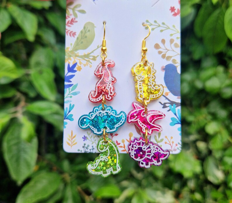 May include: Colorful dinosaur earrings with gold-colored hooks. Each earring features three dinosaur shapes in red, blue, green, yellow, pink, and purple with glitter accents. The earrings hang from a white card with floral designs.