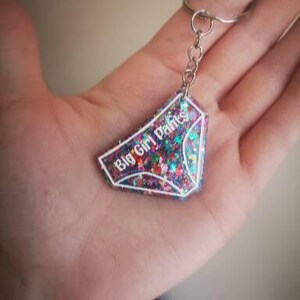 May include: A pink, purple and blue glitter keychain with a white outline in the shape of underwear. The keychain has the text "Big Girl Pants" written on it.