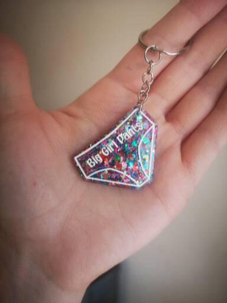 May include: A pink, purple and blue glitter keychain with a white outline in the shape of underwear. The keychain has the text "Big Girl Pants" written on it.