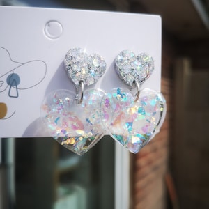 Puede incluir: Un par de pendientes de corazón plateados con purpurina iridiscente. Los pendientes tienen un pequeño dije de corazón que cuelga de la parte inferior.
