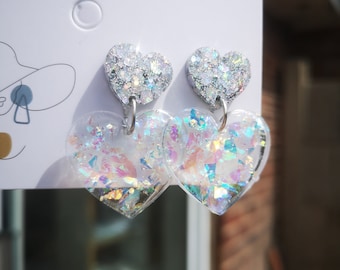 May include: A pair of silver heart-shaped earrings with iridescent glitter. The earrings have a smaller heart-shaped charm dangling from the bottom.