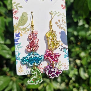 May include: Dinosaur-shaped dangle earrings with gold-colored hooks. Each earring features four colorful dinosaur shapes in red, teal, yellow, pink, and green, with glitter accents and white outlines. The earrings are displayed on a decorative card.