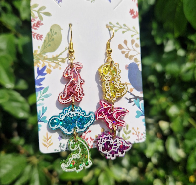 May include: Dinosaur-shaped dangle earrings with gold-colored hooks. Each earring features four colorful dinosaur shapes in red, teal, yellow, pink, and green, with glitter accents and white outlines. The earrings are displayed on a decorative card.