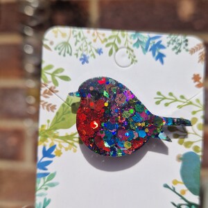 Peut inclure: Une broche de rouge-gorge scintillante et color&eacute;e avec des paillettes rouges, bleues, vertes et dor&eacute;es. La broche est sur une carte blanche avec un motif floral.