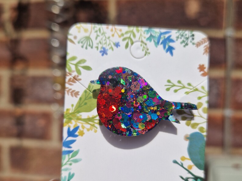 Peut inclure: Une broche de rouge-gorge scintillante et color&eacute;e avec des paillettes rouges, bleues, vertes et dor&eacute;es. La broche est sur une carte blanche avec un motif floral.