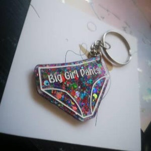 May include: A colourful glitter keychain shaped like underwear with the text "Big Girl Pants" in white lettering.