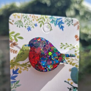 Peut inclure: Une broche en forme de rouge-gorge, recouverte de paillettes color&eacute;es, rouge, bleu, vert et violet. La broche est sur une carte blanche avec un motif floral.