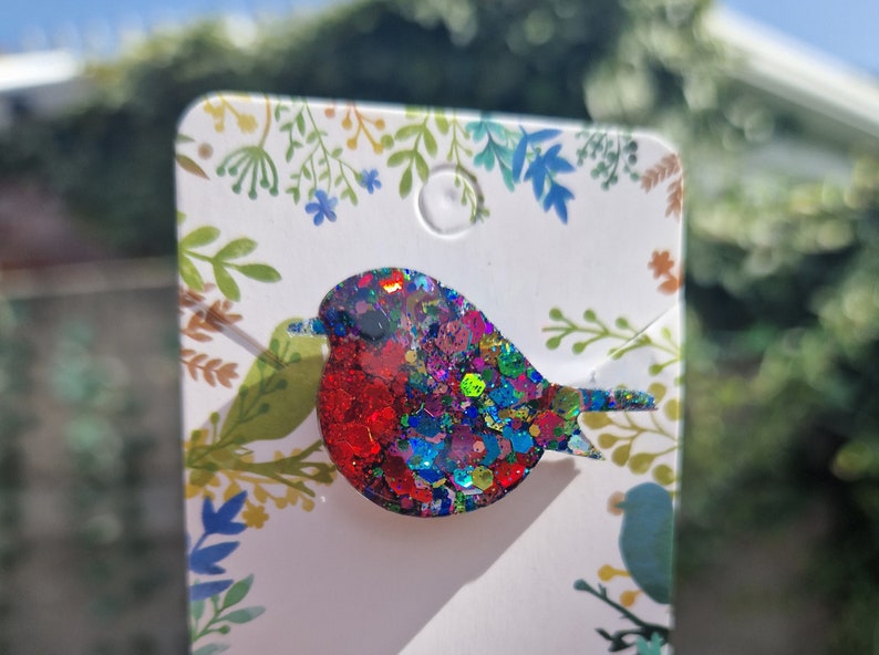 Peut inclure: Une broche en forme de rouge-gorge, recouverte de paillettes color&eacute;es, rouge, bleu, vert et violet. La broche est sur une carte blanche avec un motif floral.
