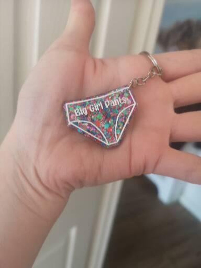 May include: A colourful glitter keychain shaped like underwear with the text "Big Girl Pants" in white.