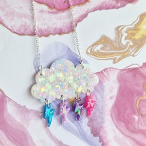 May include: A silver chain necklace featuring a cloud pendant with iridescent glitter. The cloud has four colorful lightning bolt charms in blue, purple, and pink. The necklace is displayed on a pink and purple background.