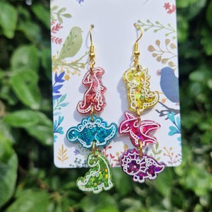 May include: Dangle earrings featuring colorful dinosaur shapes in red, blue, green, yellow, and purple. The earrings have gold-colored hooks and are displayed on a white card with floral and bird illustrations. The dinosaurs have glitter accents.