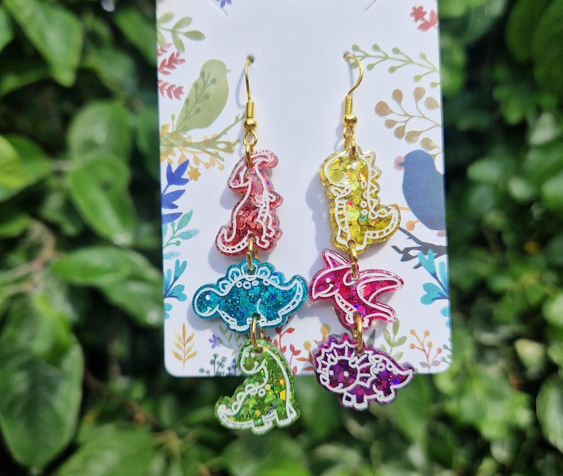 May include: Dangle earrings featuring colorful dinosaur shapes in red, blue, green, yellow, and purple. The earrings have gold-colored hooks and are displayed on a white card with floral and bird illustrations. The dinosaurs have glitter accents.