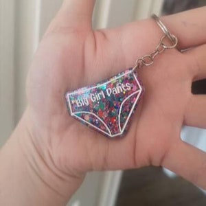 May include: A pink and white glitter keychain shaped like underwear with the text "Big Girl Pants" in white.