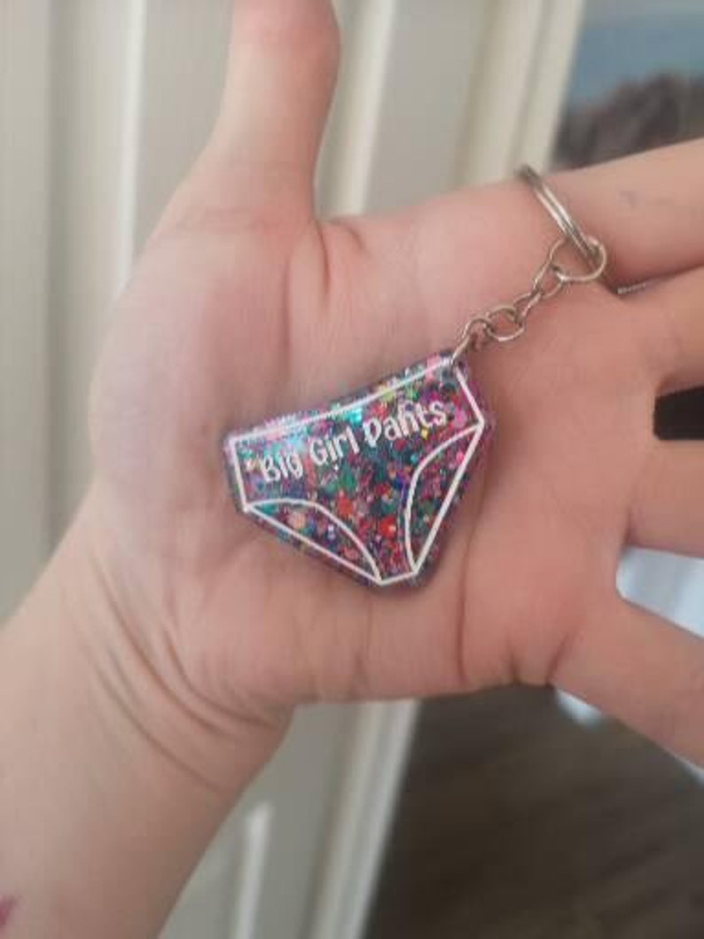 May include: A pink and white glitter keychain shaped like underwear with the text "Big Girl Pants" in white.