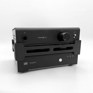 May include: A black Schiit Magni headphone amplifier stacked on top of a Schiit Modi digital-to-analogue converter, both held together by a black 3D-printed plastic bracket. The Magni features a volume knob, headphone jack, and toggle switch.