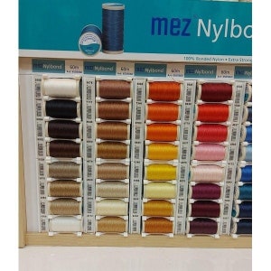 May include: A display of 60 meter spools of mez Nylbond thread in various colors. The thread is 100% bonded nylon and extra strong. The spools are arranged in rows on a wooden shelf.