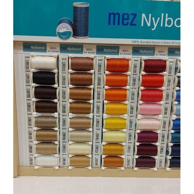 May include: A display of 60 meter spools of mez Nylbond thread in various colors. The thread is 100% bonded nylon and extra strong. The spools are arranged in rows on a wooden shelf.