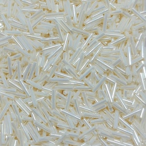 Może przedstawiać: Zbliżenie na stos białych, opalizujących szklanych koralików. Koraliki są długie i cienkie, przypominające małe cylindry.