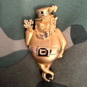 May include: A gold-colored leprechaun figurine with a green shamrock hat, holding a pipe and a shamrock in his hand. The figurine is wearing a green belt with a buckle.