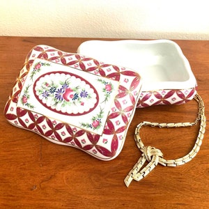 May include: A white porcelain trinket box with a red and gold floral design and a matching lid. The box is open and a gold chain necklace with a knotted pendant is lying next to it.