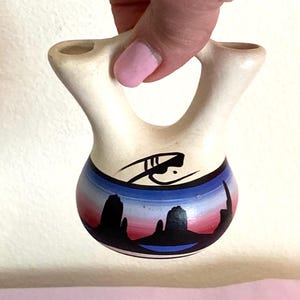 May include: A cream-colored, double-spouted ceramic vase with a handle. The vase features a painted landscape scene with red, blue, and black colors. The top of the vase has a black painted design.