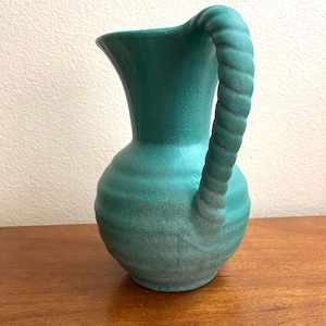 May include: A teal blue ceramic pitcher with a rope-like handle. The pitcher has a textured surface and a rounded body.