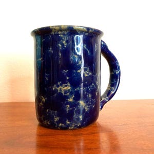 May include: A ceramic mug with a dark blue speckled glaze. The mug has a rounded handle and a slightly flared rim. The speckled pattern includes areas of light tan and cream. The mug is sitting on a wooden surface.