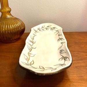 May include: Cream-colored, leaf-decorated serving dish with a gold rim. The dish has a unique, irregular shape and is adorned with hand-painted gray and gold leaf designs. A vintage amber glass bottle is in the background.