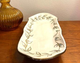 May include: Cream-colored, leaf-decorated serving dish with a gold rim. The dish has a unique, irregular shape and is adorned with hand-painted gray and gold leaf designs. A vintage amber glass bottle is in the background.