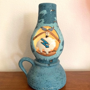 May include: A teal ceramic lamp with a handle and a round base. The lamp features a cutout with a white pig wearing sunglasses and a blue lightning bolt. The lamp has a textured surface and a warm glow.