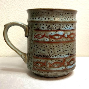 May include: A ceramic mug with a speckled teal and brown glaze. The mug features horizontal bands of light blue with a repeating fish design in a reddish-brown color. The handle is a matching teal color.