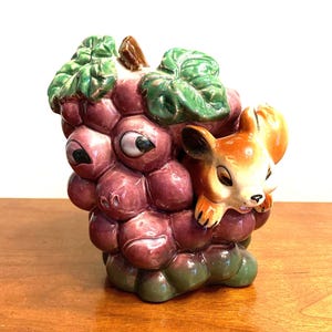 May include: A ceramic figurine of a purple grape cluster with a brown and white squirrel peeking out from behind the grapes.