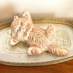 May include: A ceramic figurine of a light brown cat lying on its back with its paws in the air. The cat has blue eyes and pink accents on its paws and ears. The figurine is on a decorative silver tray with a gold rim.