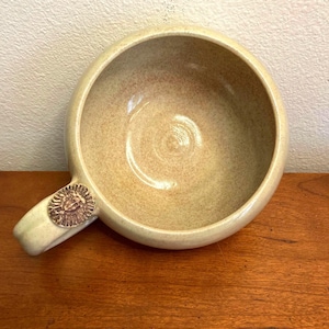 May include: A large, beige ceramic soup bowl with a rounded shape and a handle. The handle has a raised, brown design.