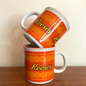 May include: Two orange and white ceramic mugs stacked on top of each other. The mugs feature the Reese's logo in yellow and orange, with a pattern of orange circles and stripes. Each mug has a white handle.