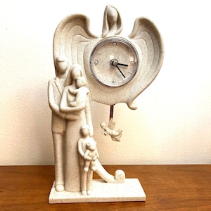 May include: A white ceramic clock with a pendulum featuring a family of four and a winged angel. The clock face is round and has black hands and Roman numerals.