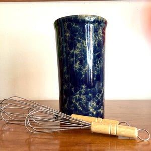 May include: A tall, dark blue ceramic utensil holder with a speckled pattern, paired with two stainless steel whisks with wooden handles. The holder is cylindrical and stands upright on a wooden surface.