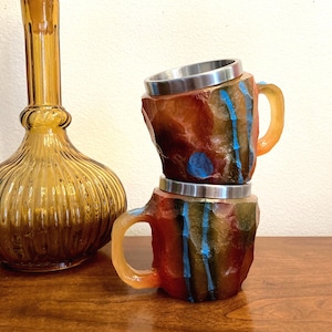 May include: Two stacked ceramic mugs with a rock-like texture. The mugs are painted with brown, orange, green, and blue stripes. The mugs have a stainless steel rim and a handle.