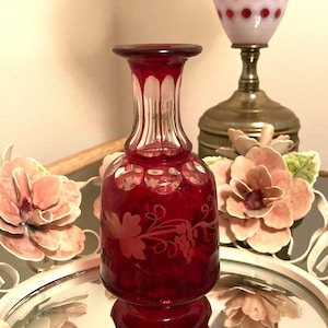 Puede incluir: Una pequeña botella de vidrio roja con un tapón de vidrio transparente. La botella tiene un diseño grabado en blanco de una vid con hojas y uvas. La botella está sobre una superficie de espejo.