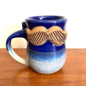 May include: A handmade ceramic mug with a blue glaze that fades from dark to light. A tan-colored mustache is affixed to the front of the mug. The handle is blue with a white accent. The mug is sitting on a wooden surface.