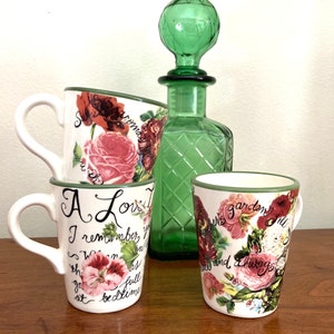 May include: Three white ceramic mugs with floral designs and green rims. The mugs have handwritten text that reads "A Love I remember you", "Sweet romance with", and "In this garden full of", respectively.