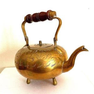 May include: A vintage brass teapot with a round body and a curved spout. The teapot features a wooden handle and is supported by three small legs. The surface is decorated with floral engravings, adding a decorative touch to the piece.