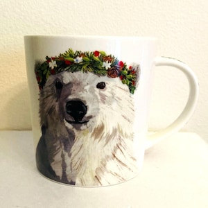 May include: White ceramic mug featuring a watercolor illustration of a polar bear wearing a floral wreath. The bear has a black nose and eyes, with a fur texture. The mug has a curved handle and a white background.