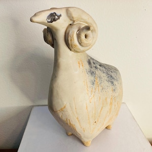 May include: A ceramic ram sculpture with a cream-colored body, large curled horns, and a dark spot near the eye. The ram has a textured surface with hints of blue and orange, and stands on three small legs.