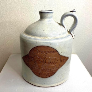 May include: A ceramic jug with a speckled off-white glaze and a single handle. The front features a brown, diamond-shaped medallion embossed with the text Oregon Trail, a mountain range, a covered wagon, and the years 1843 and 1975.